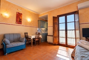 A room with a sofa, dining table, chairs, a kitchen area, and a balcony with curtains. - Case Vacanze I Chiari - 40 Camera doppia (Montepulciano)