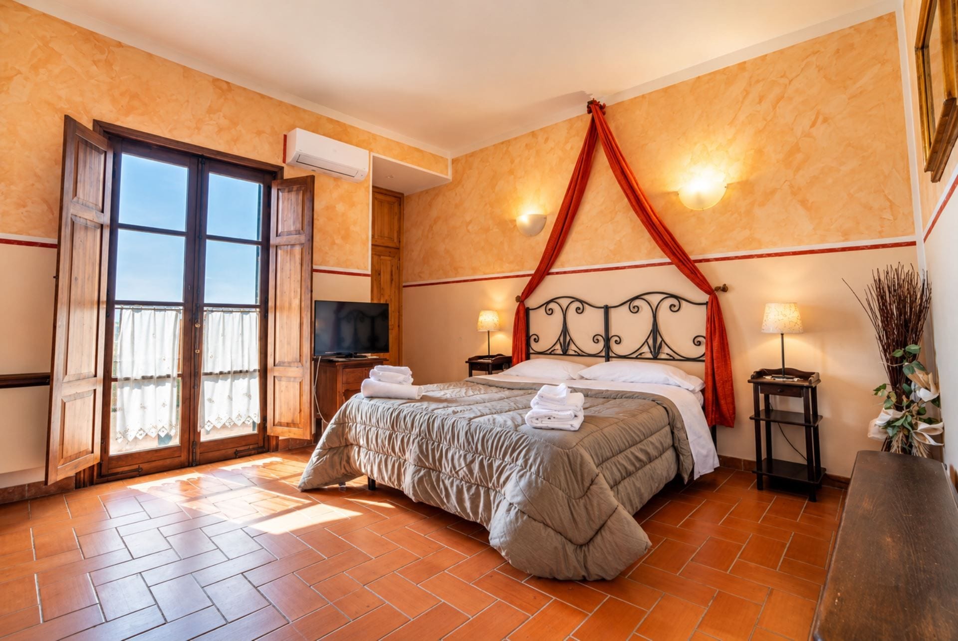 Un dormitorio con cama, televisión, ventana con cortinas, aire acondicionado montado en la pared y lámpara de noche.