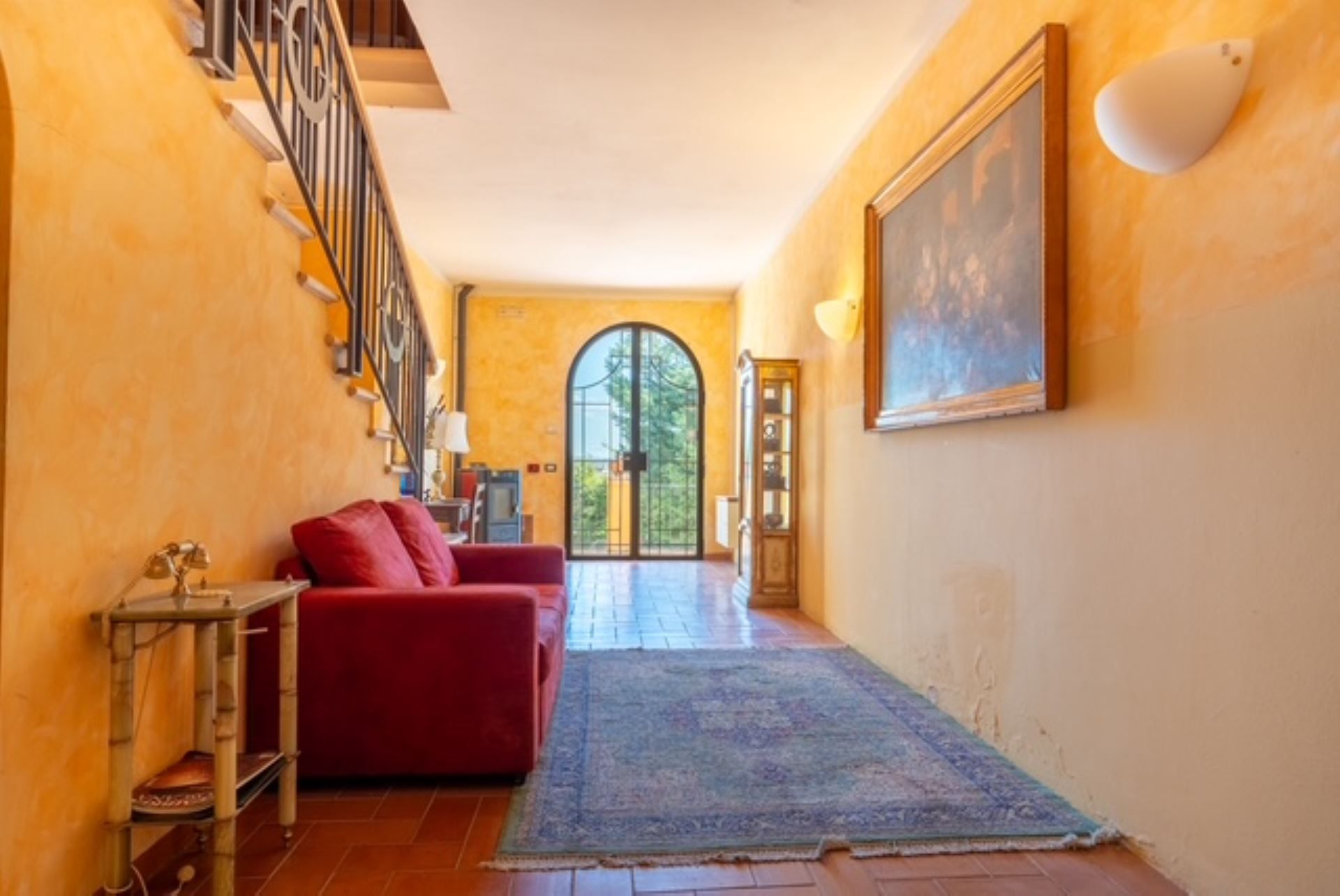 Un couloir avec un canapé rouge, une petite table, un tapis bleu, un escalier et un grand tableau accroché au mur.