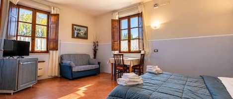 Habitación de hotel con cama, sofá, escritorio con sillas, televisión y ventana con cortinas.