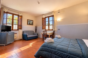 Una camera d'albergo con un letto, un divano, una scrivania con sedie, una televisione e una finestra con tende.