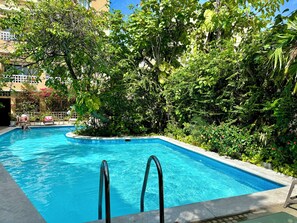 A swimming pool surrounded by lush greenery and a building in the background. - by Calamari.Homes - Centenario Casa 102 - Old City (Cartagena de Indias)