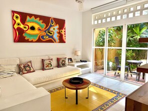 A living room with a white sofa, colorful cushions, a large abstract painting, a small table with a bowl, and a view of a garden through a glass door. - by Calamari.Homes - Centenario Casa 102 - Old City (Cartagena de Indias)