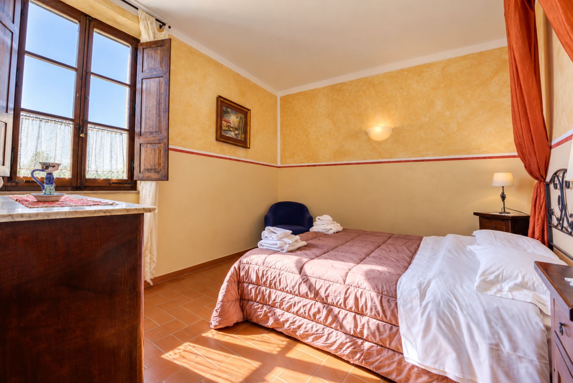 Habitación de hotel con cama, una silla, un escritorio con un jarrón y un cuadro en la pared.