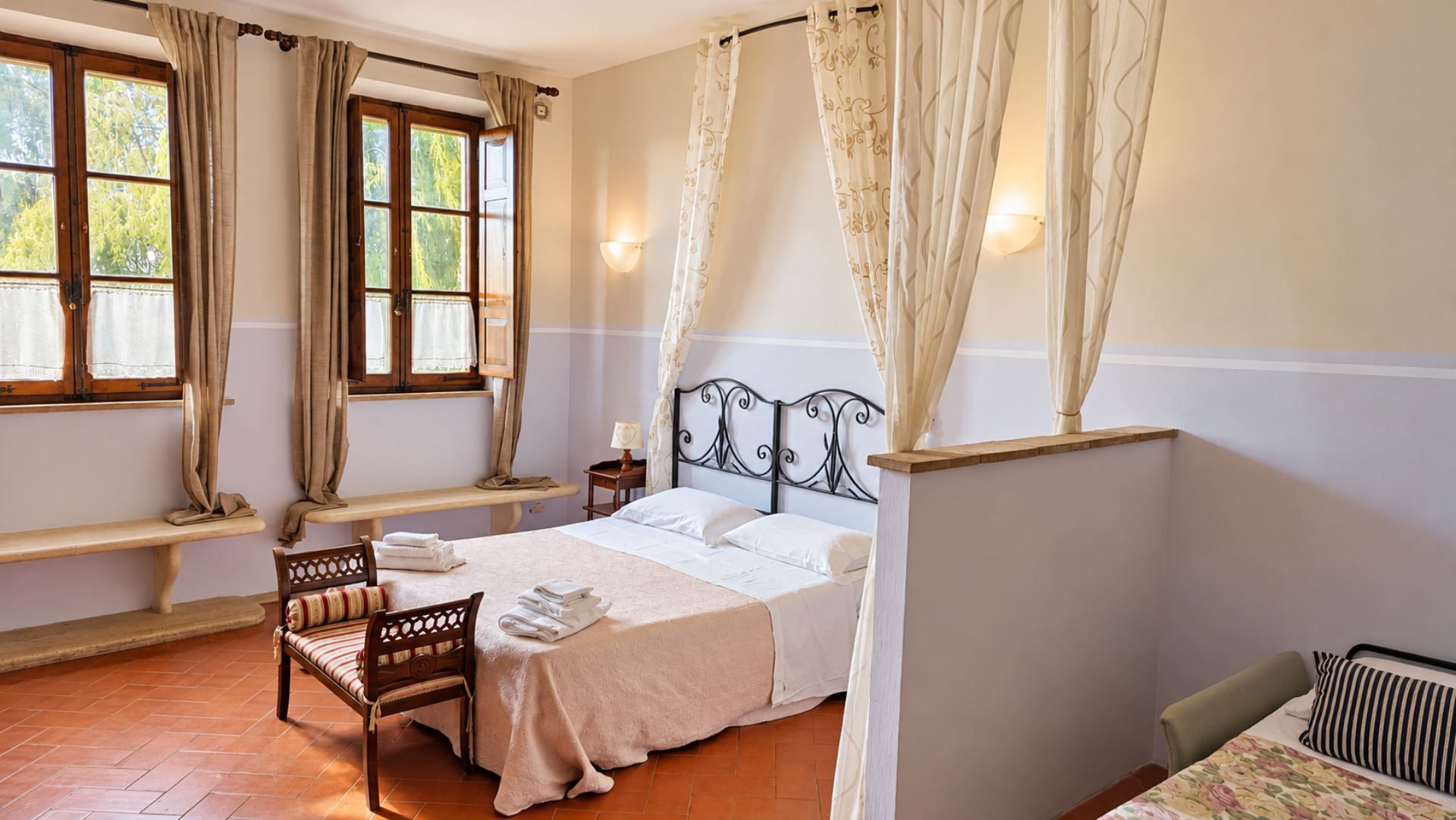 Un dormitorio con cama, una banca, una silla, una mesita de noche y una ventana con cortinas.