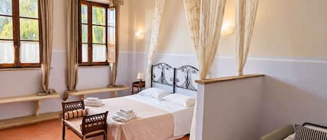 Un dormitorio con cama, una banca, una silla, una mesita de noche y una ventana con cortinas.
