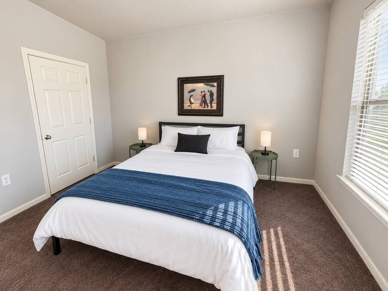 A bedroom with a bed, a door, a framed picture on the wall, two bedside tables with lamps, and a window with blinds.