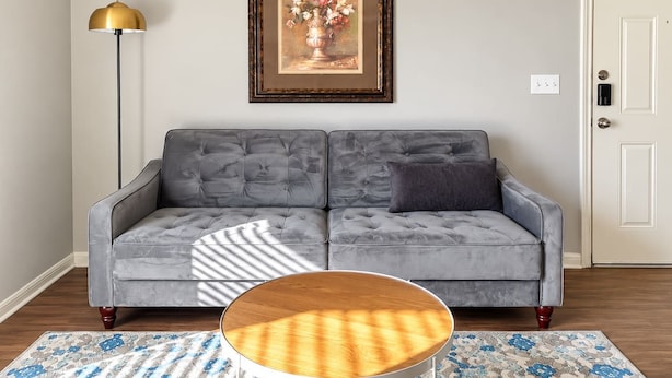 Ein graues, gepolstertes Sofa mit einem dunklen Kissen, ein runder Couchtisch aus Holz, ein gerahmtes Blumenbild, eine Stehlampe und eine Tür mit Schloss.