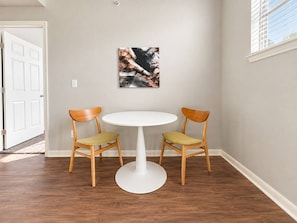 A dining area with a round table and two chairs, a painting on the wall, and a door in the background. - Sleepover | Fresh 1BD/1BA + Gym - Corpus Christi (Corpus Christi)