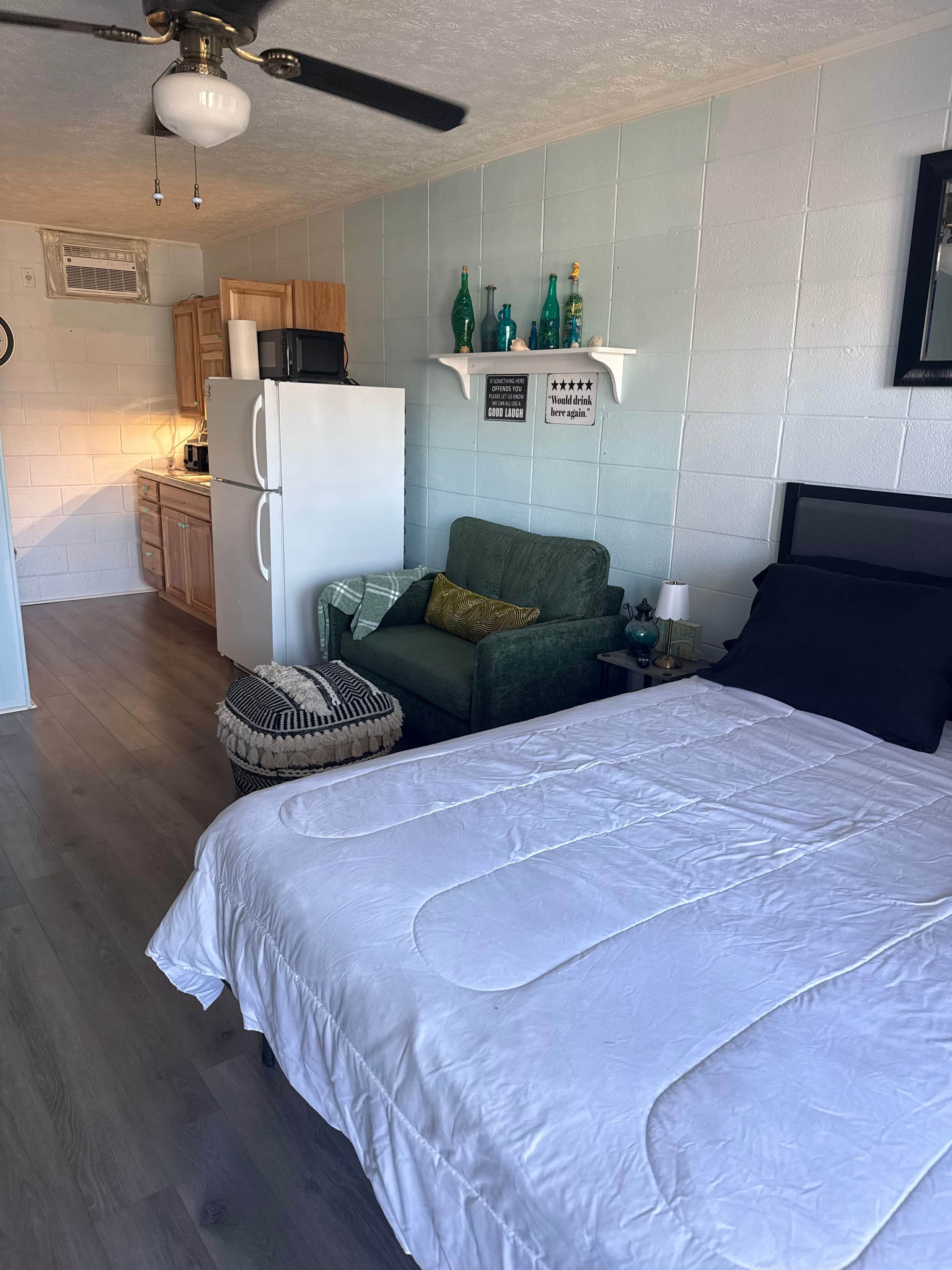 Une chambre à coucher avec un lit, un canapé vert, une petite table avec une lampe, et, au fond, une kitchenette.