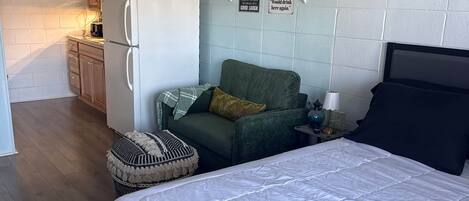 A bedroom with a bed, a green sofa, a small table with a lamp, and a kitchenette in the background.
