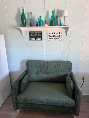 A green sofa in a room with a white brick wall, a shelf with decorative bottles, and two signs with messages. - Oceans 11, Vegas-style Studio, pet/ok steps to bch (North Myrtle Beach)