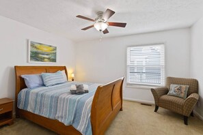 Una camera da letto con un letto, una sedia, una ventola a soffitto, una finestra con persiane e un quadro appeso al muro.