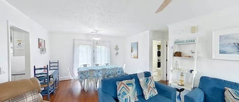 A living room with a blue sofa set, a dining table with chairs, a ceiling fan, and a kitchen area in the background.