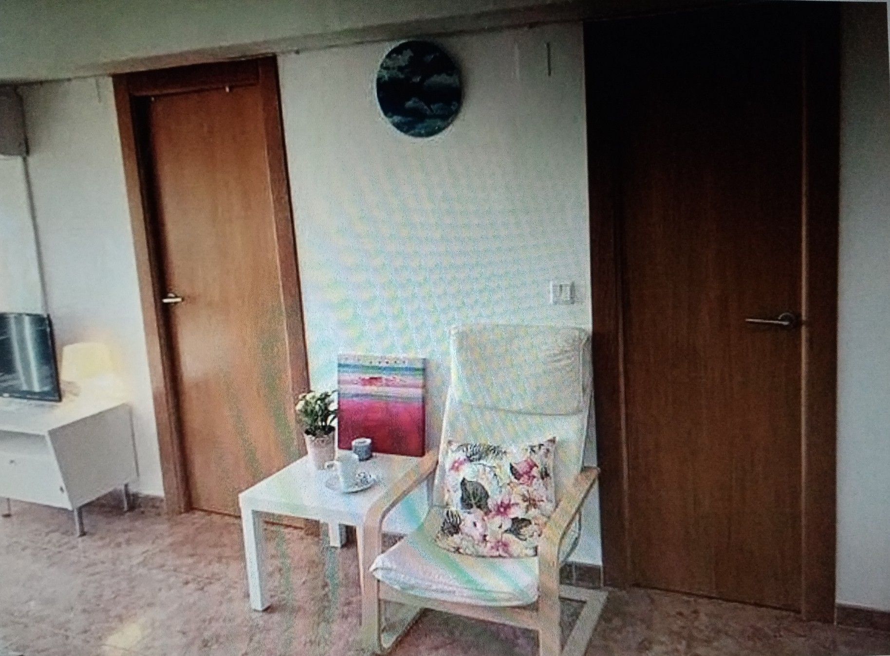 Una habitación con puerta de madera, una mesita con una planta, una silla con cojín floral y un objeto redondo sujeto a la pared.