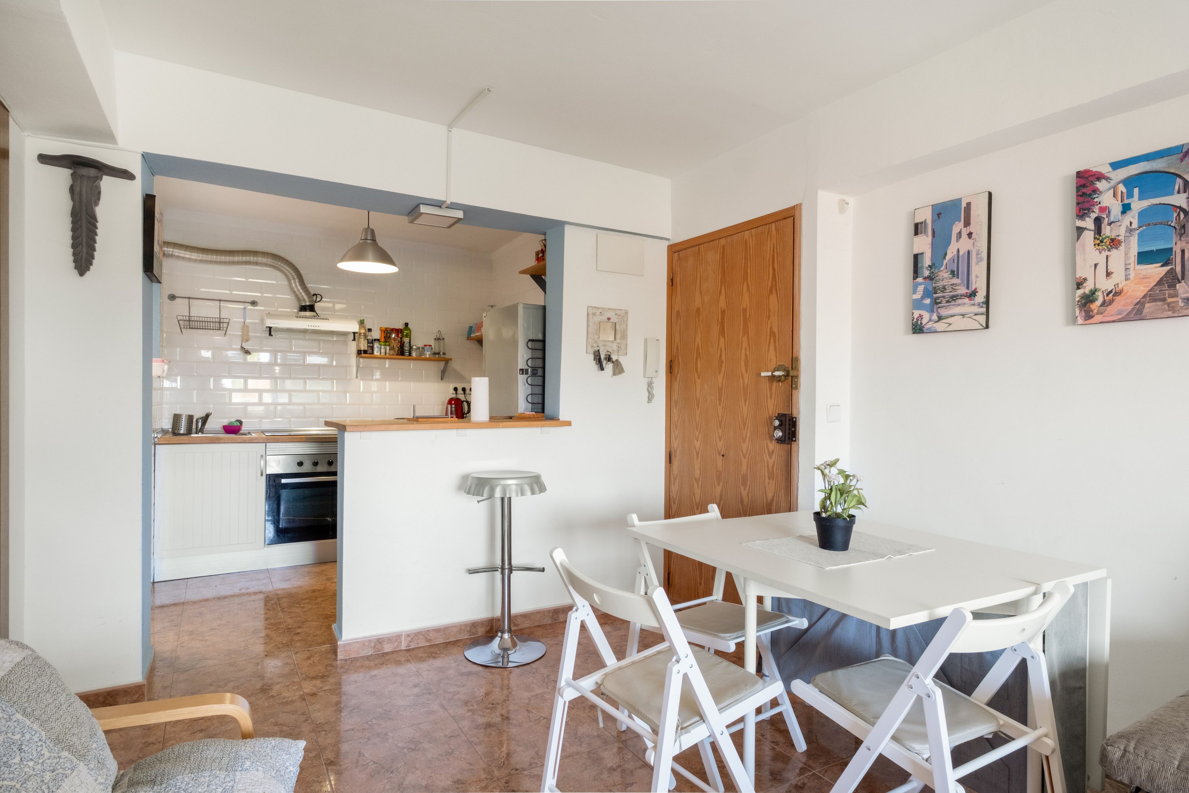 Een compacte keuken met witte kasten, een inbouwoven en een houten deur naar een eethoek met een witte tafel en stoelen.