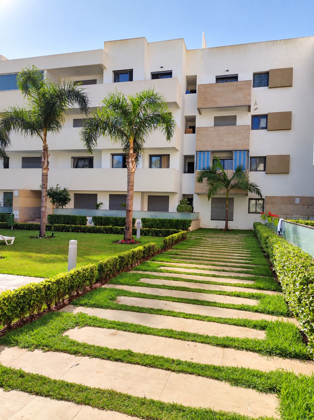 A modern multi-story residential building with a central courtyard featuring a patterned walkway, palm trees, and well-maintained greenery.