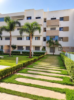 A modern multi-story residential building with a central courtyard featuring a patterned walkway, palm trees, and well-maintained greenery. - Beautiful 2-bedroom Apartment With Pool View Mohammedia (El Mansouria)