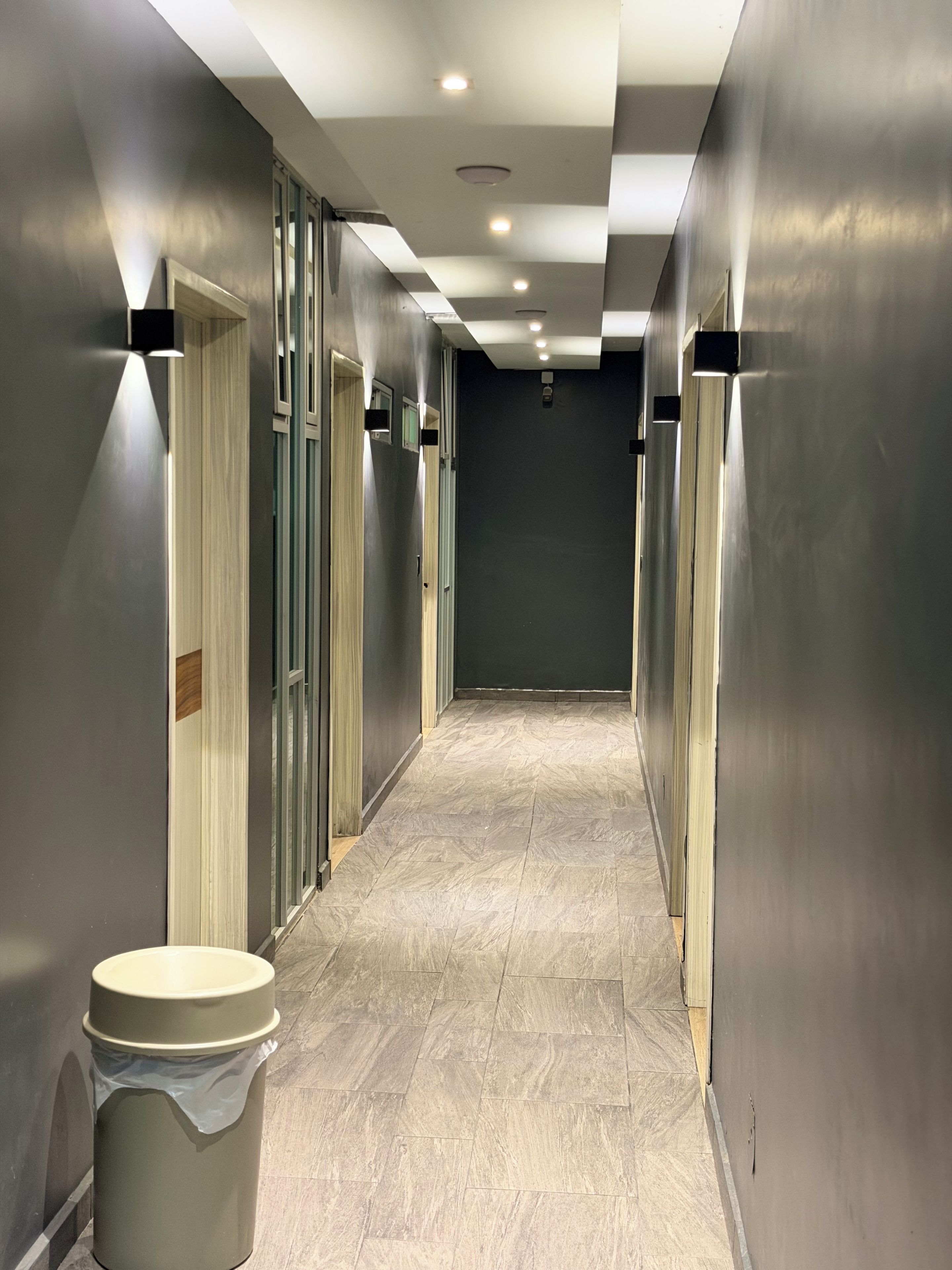 A hallway with doors on both sides, a trash bin in the foreground, and a tiled floor.