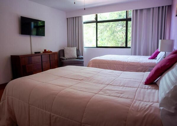 A hotel room with two beds, a TV mounted on the wall, a wooden dresser, and a window with curtains.