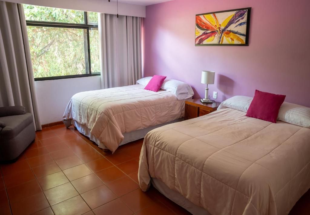A hotel room with two beds, a window with curtains, a chair, and a painting on the wall.