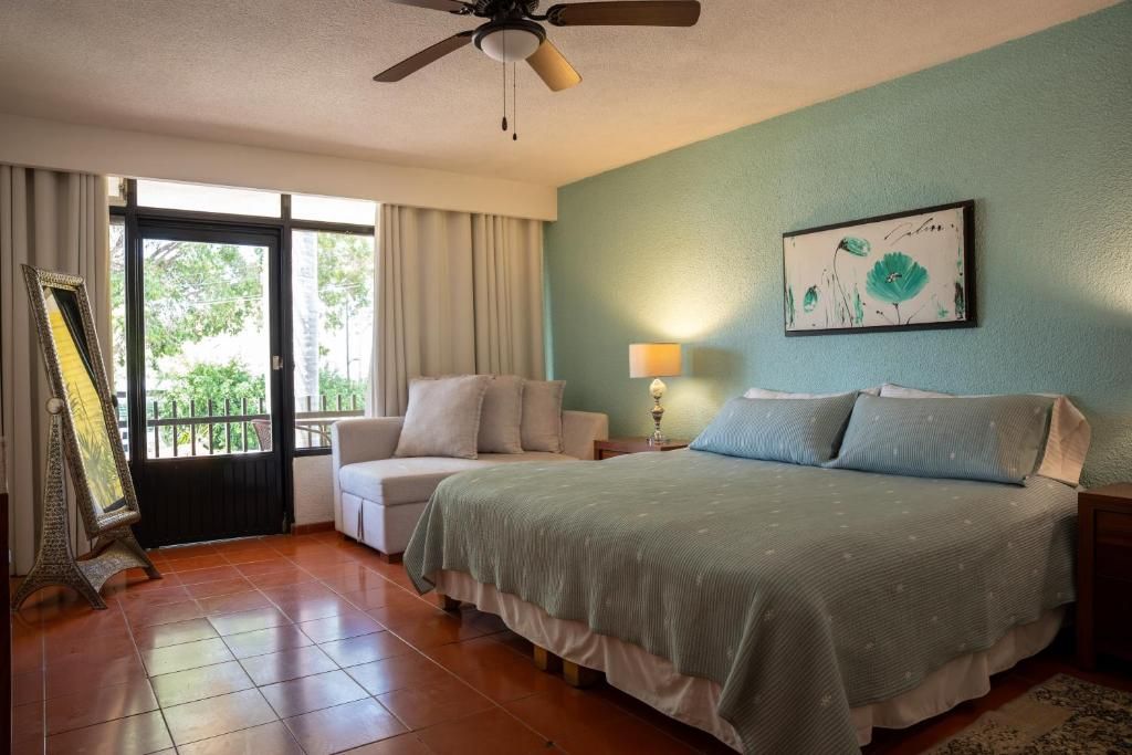 Habitación de hotel con cama, silla, mesita de noche, lámpara, ventilador de techo y ventana con cortinas.