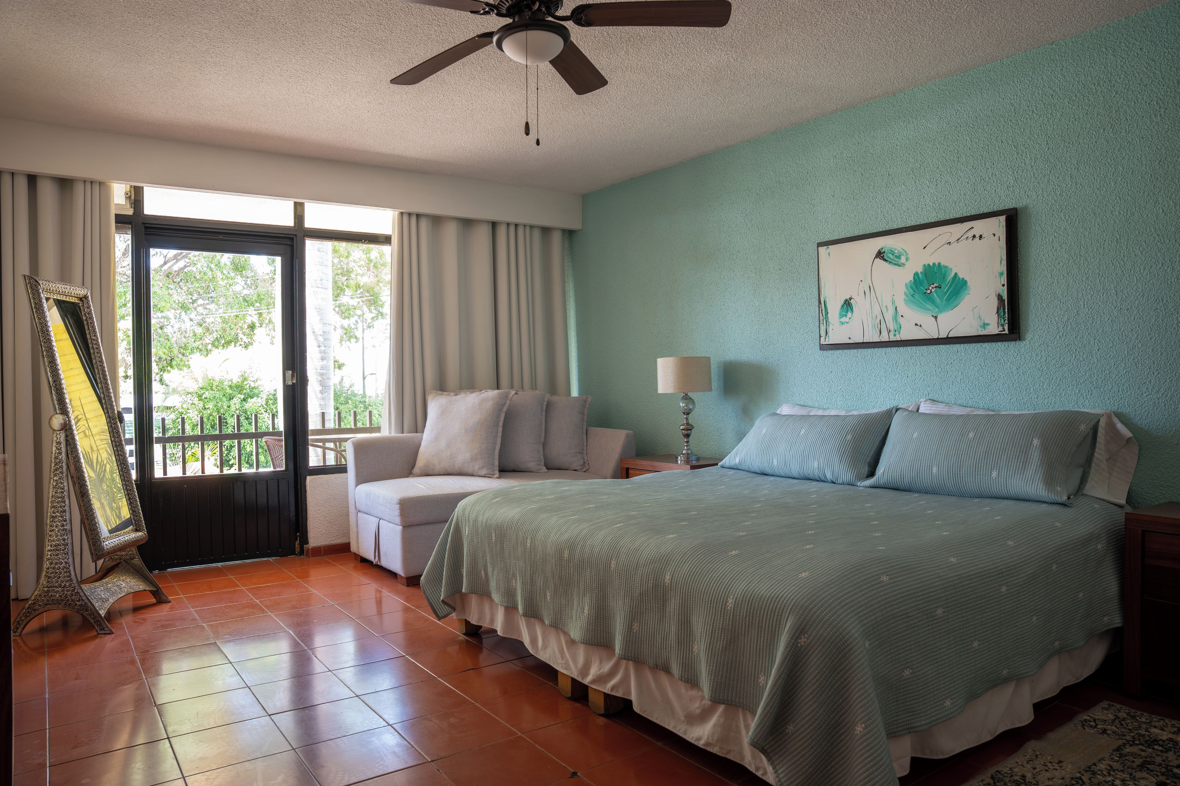 Un dormitorio con cama, una silla, mesita de noche, lámpara, ventilador de techo y ventana con cortinas.