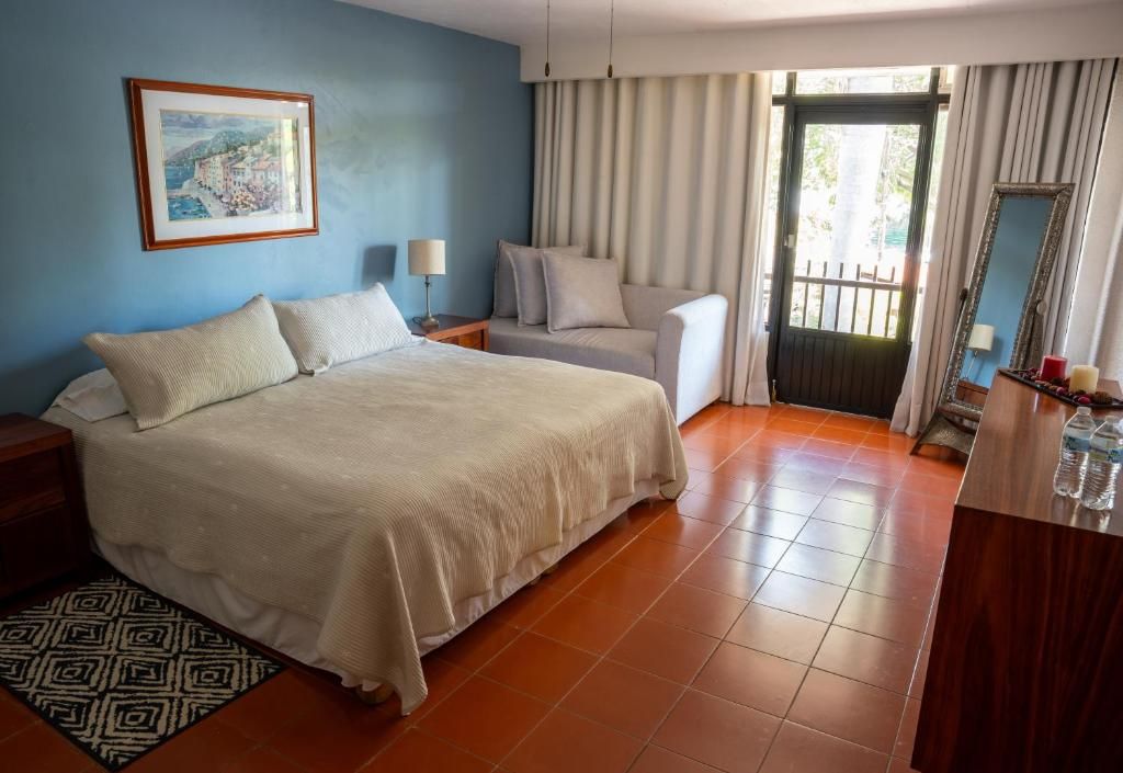A hotel room with a bed, a chair, a desk, a mirror, a painting, and a door with curtains.