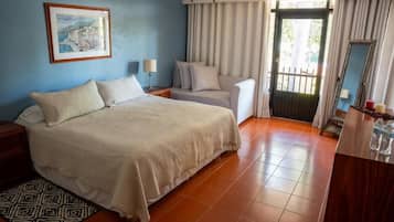 A hotel room with a bed, a chair, a desk, a mirror, a painting, and a door with curtains.