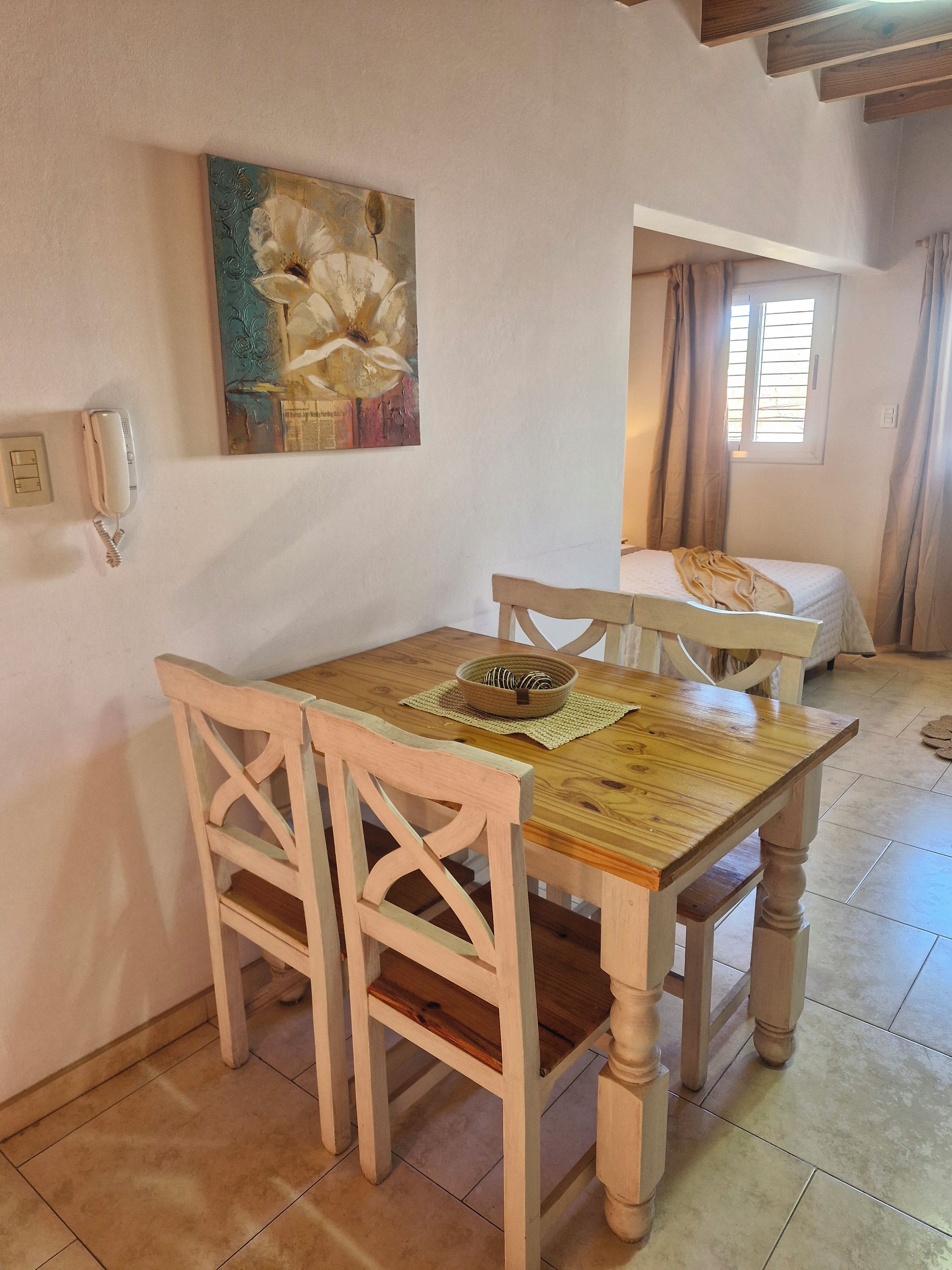 Una habitación con una mesa de comedor de madera, dos sillas, una cama, una ventana con cortinas y un cuadro en la pared.