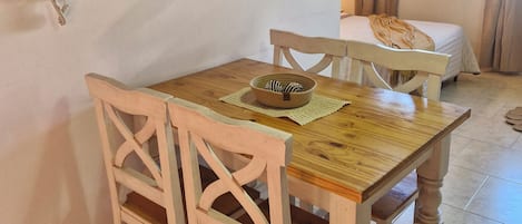 A room with a wooden dining table, two chairs, a bed, a window with curtains, and a painting on the wall.