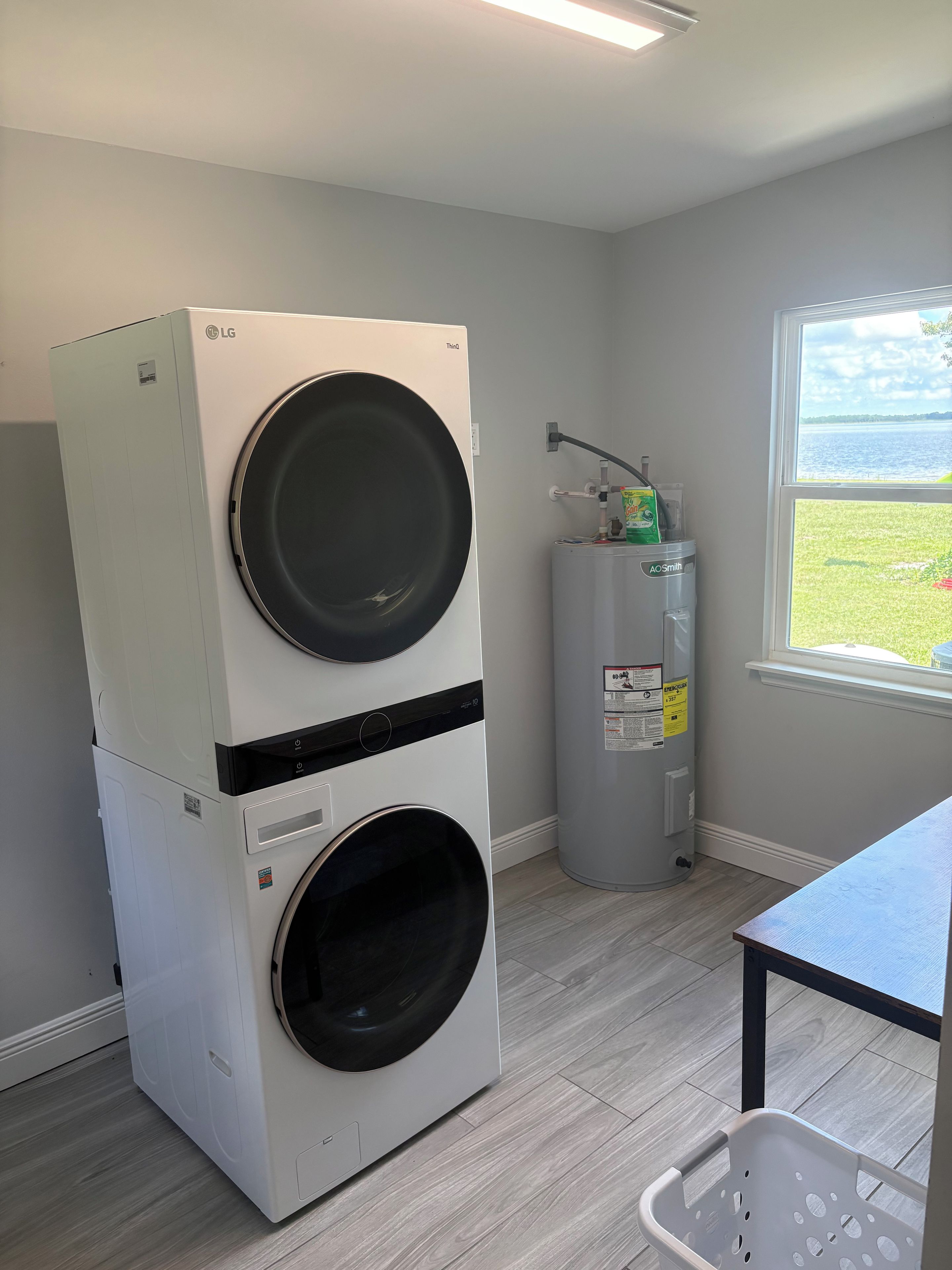 A laundry room with stacked washer and dryer units, a water heater, and a window with a view of a grassy area.