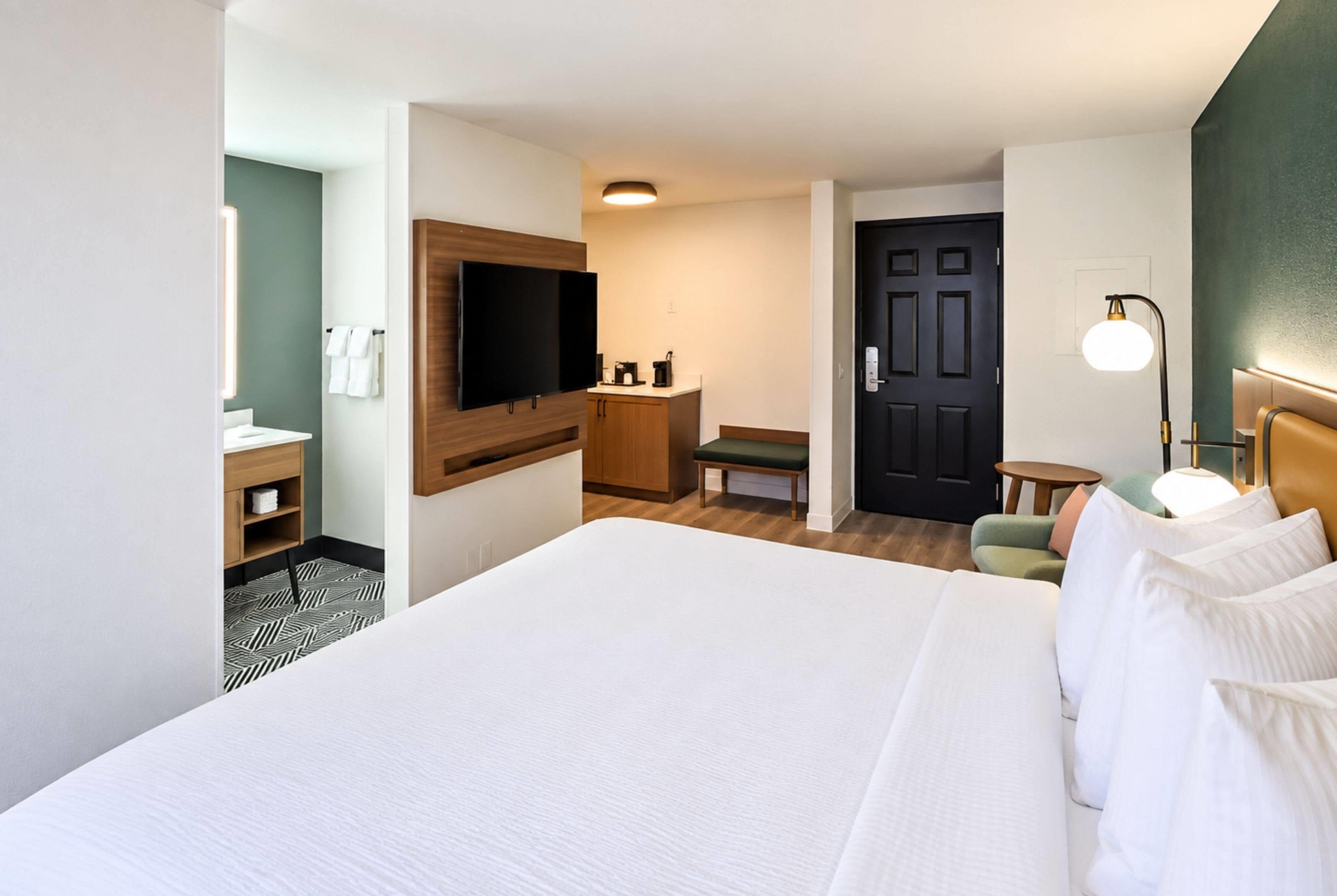 Une chambre d’hôtel avec un grand lit, un téléviseur fixé au mur, un bureau avec une chaise, une petite table et une porte.