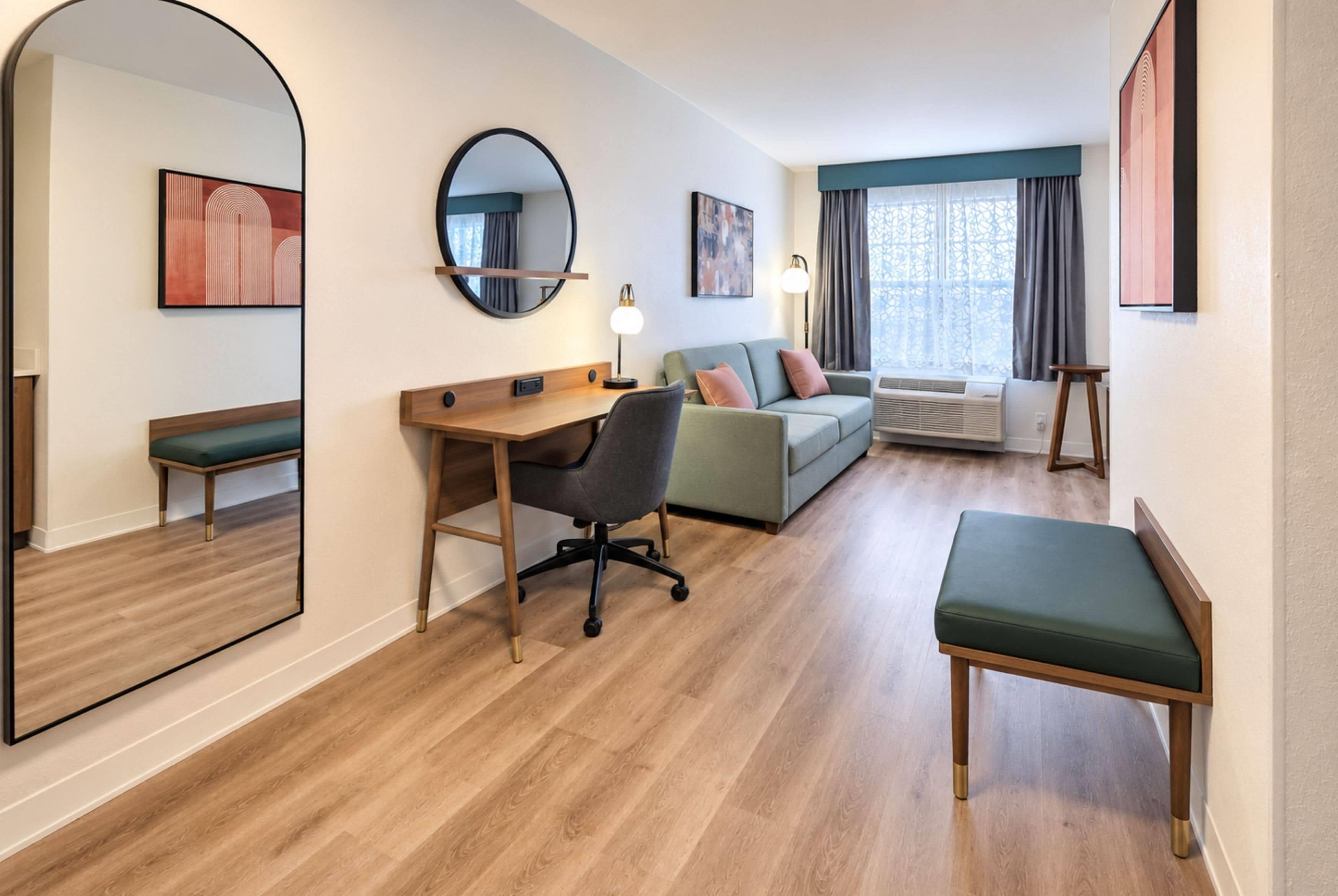 Et hotelværelse med trægulv, et skrivebord med stol, en sofa, en bænk, et stort spejl og billedkunst på væggene.