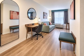 Une chambre d’hôtel avec un plancher en bois, un bureau avec une chaise, un canapé, un banc, un grand miroir et des œuvres d’art accrochées aux murs.