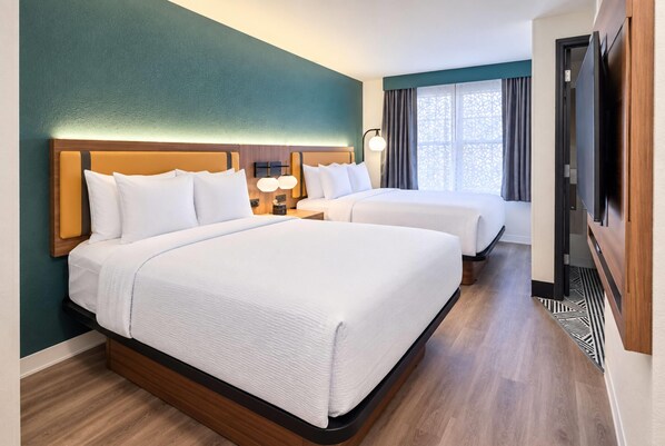 Habitación de hotel con dos camas, cabecera de madera, mesita de noche con lámpara y ventana con cortinas.