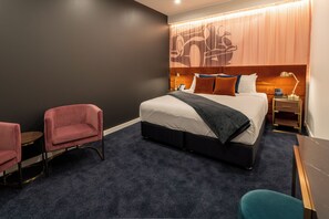 Habitación de hotel con cama, dos sillas, escritorio, mesita de noche y pared con diseño decorativo.