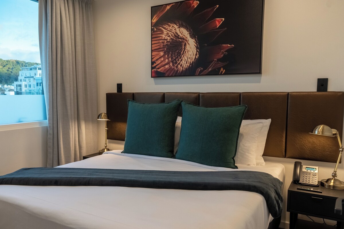 Una habitación de hotel con una cama, dos almohadas, una mesita de noche con un teléfono, una lámpara y un cuadro en la pared.