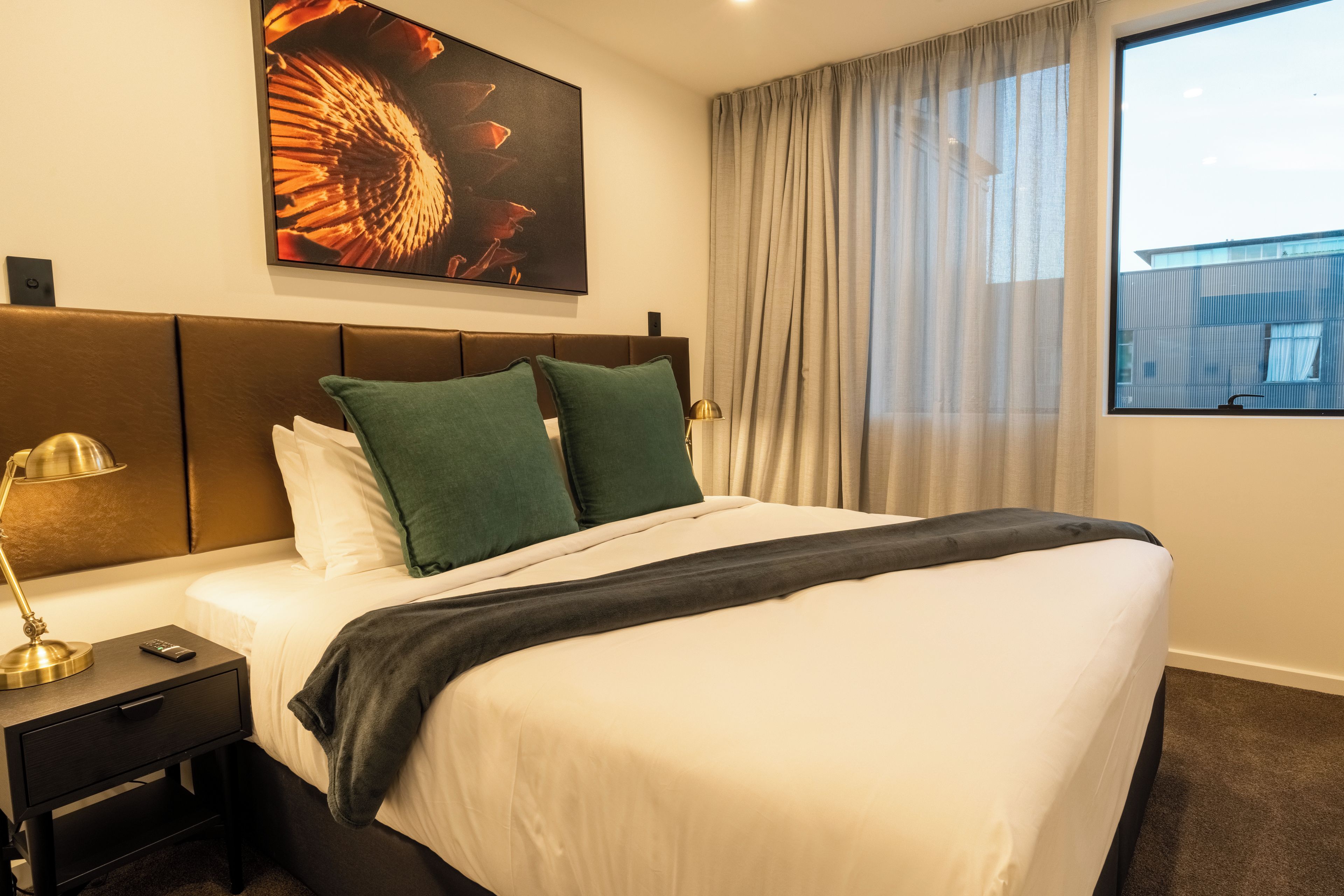 Una habitación de hotel con una cama, una mesita de noche con una lámpara, un cuadro en la pared y una ventana con cortinas.