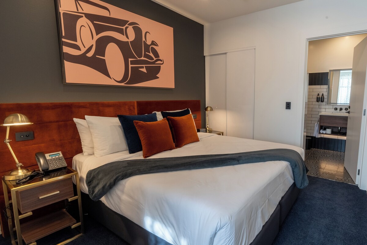 Habitación de hotel con una cama grande, una mesita de noche con lámpara, una obra de arte en la pared y vistas al baño.