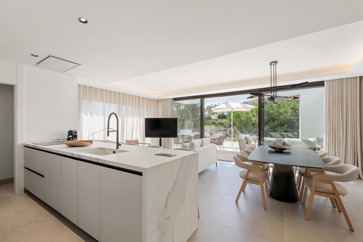 A modern kitchen with a large island, a dining area with a round table, and a view of the outdoors through large windows.