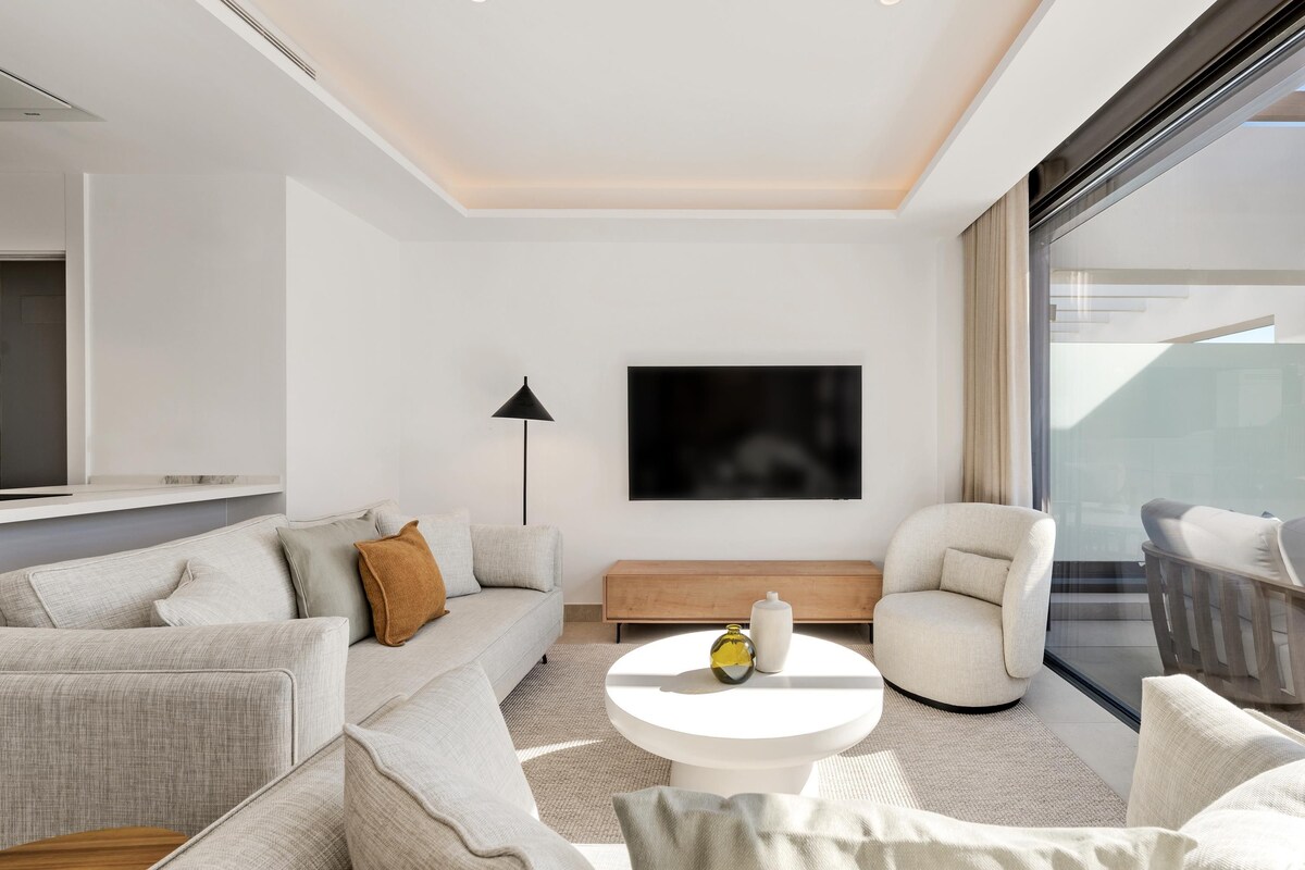 A modern living room with a sofa, armchair, coffee table, and a flat-screen TV mounted on the wall.