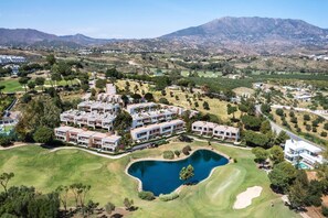 Un campo de golf con un lago, edificios modernos y montañas al fondo.