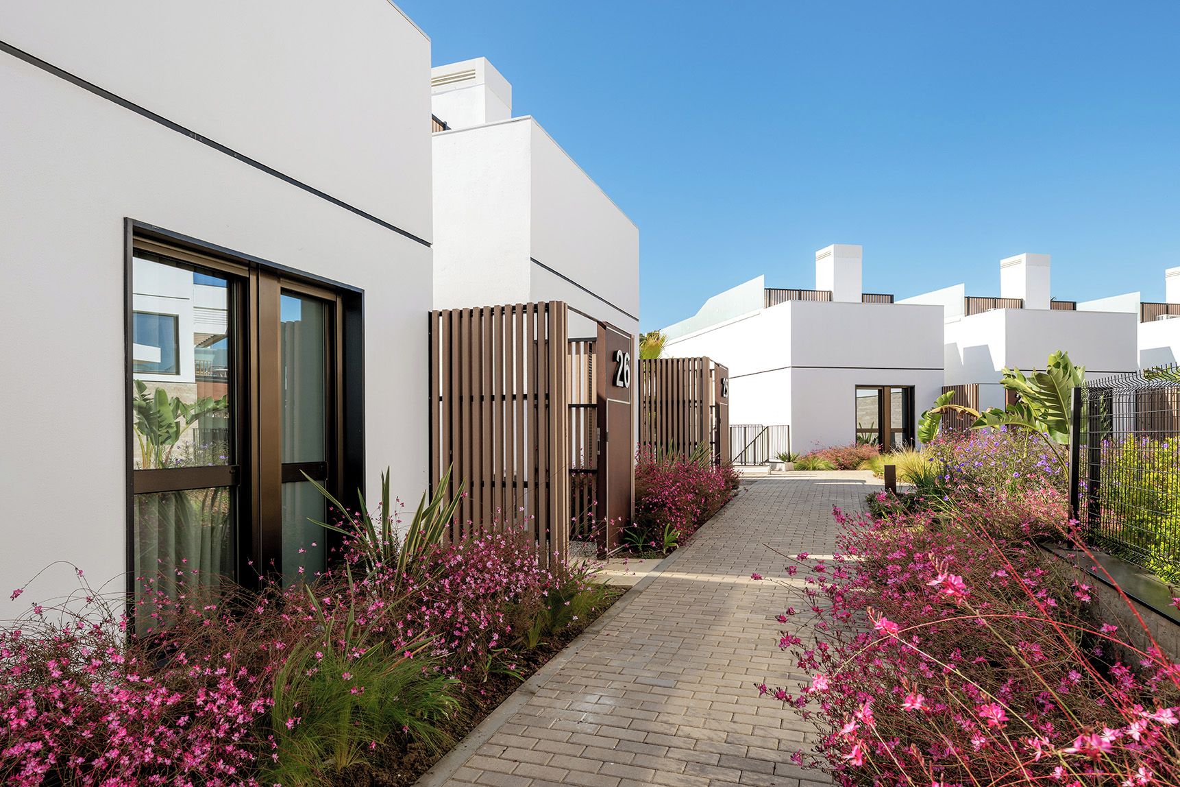 A modern building with a brick pathway, wooden fence, and pink flowering plants.