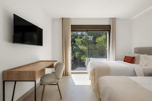 A modern hotel room with a flat-screen TV, a wooden desk, a comfortable chair, a large bed with pillows, and a balcony with a view of trees.