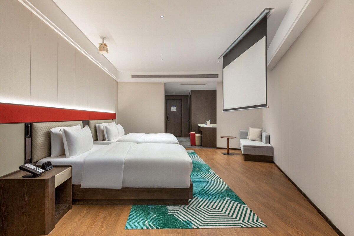 Une chambre d’hôtel comprenant un lit, des tables de chevet, un bureau, une chaise, une petite table, un canapé et un écran de projection.