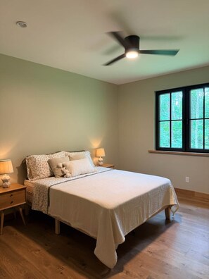 Un dormitorio con cama, mesitas de noche, ventilador de techo y ventana con vista a árboles.