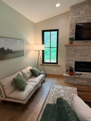 A living room with a sofa, a lamp, a fireplace, a TV, and a window with a view of greenery.