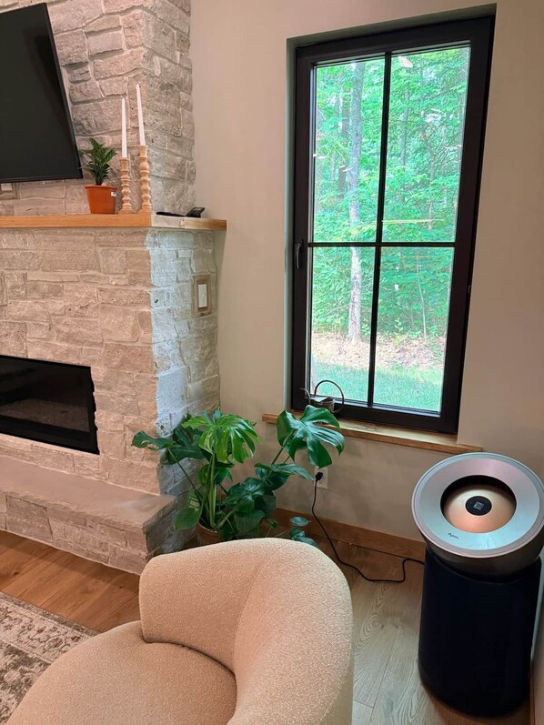 A stone fireplace with a flat-screen TV, a window with a view of trees, and a round speaker on the floor.
