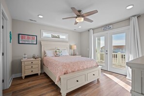 A bedroom with a bed, nightstands, a ceiling fan, and a view of the outside. - Ocean Views with Heated Pool! One Row from Beach! (Oak Island)